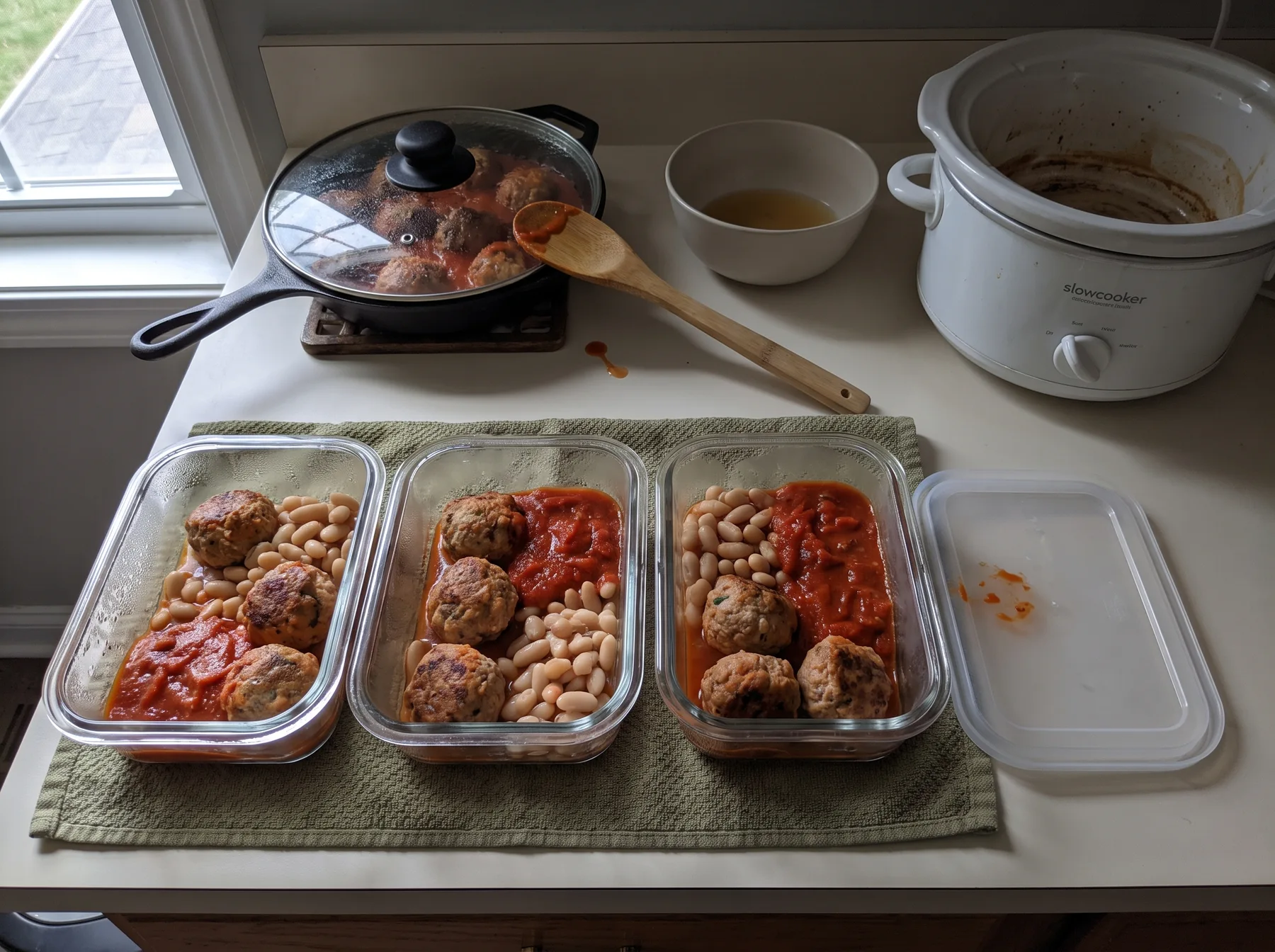 Three shallow containers hold turkey meatballs, white beans, and red pepper sauce beside a slow cooker.
