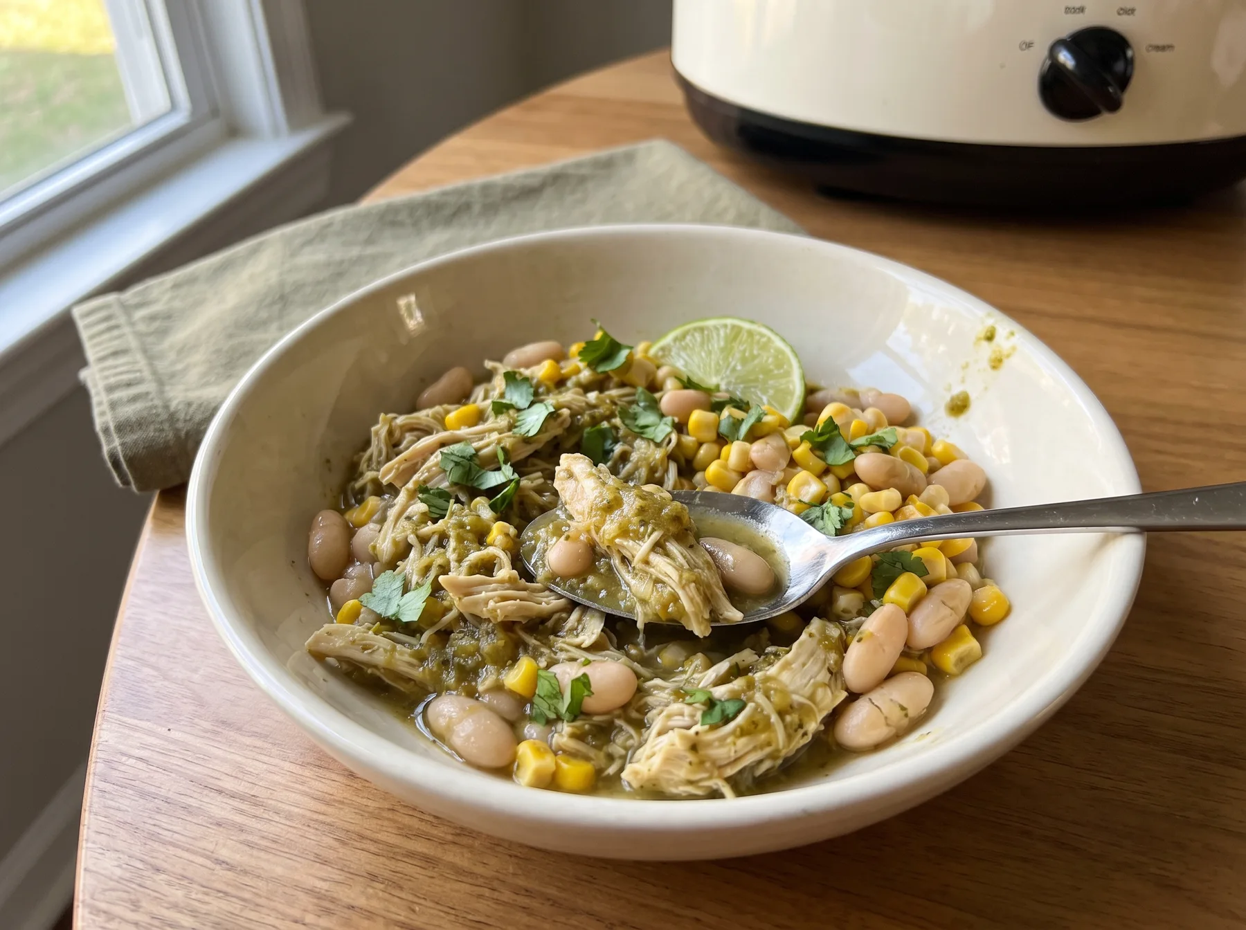 Bowl of shredded salsa verde chicken with white beans, corn, lime, and cilantro beside a slow cooker