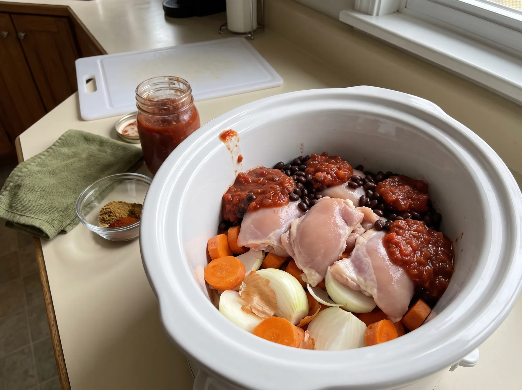 Raw chicken thighs over carrots and onion with beans and sauce in an open slow cooker.