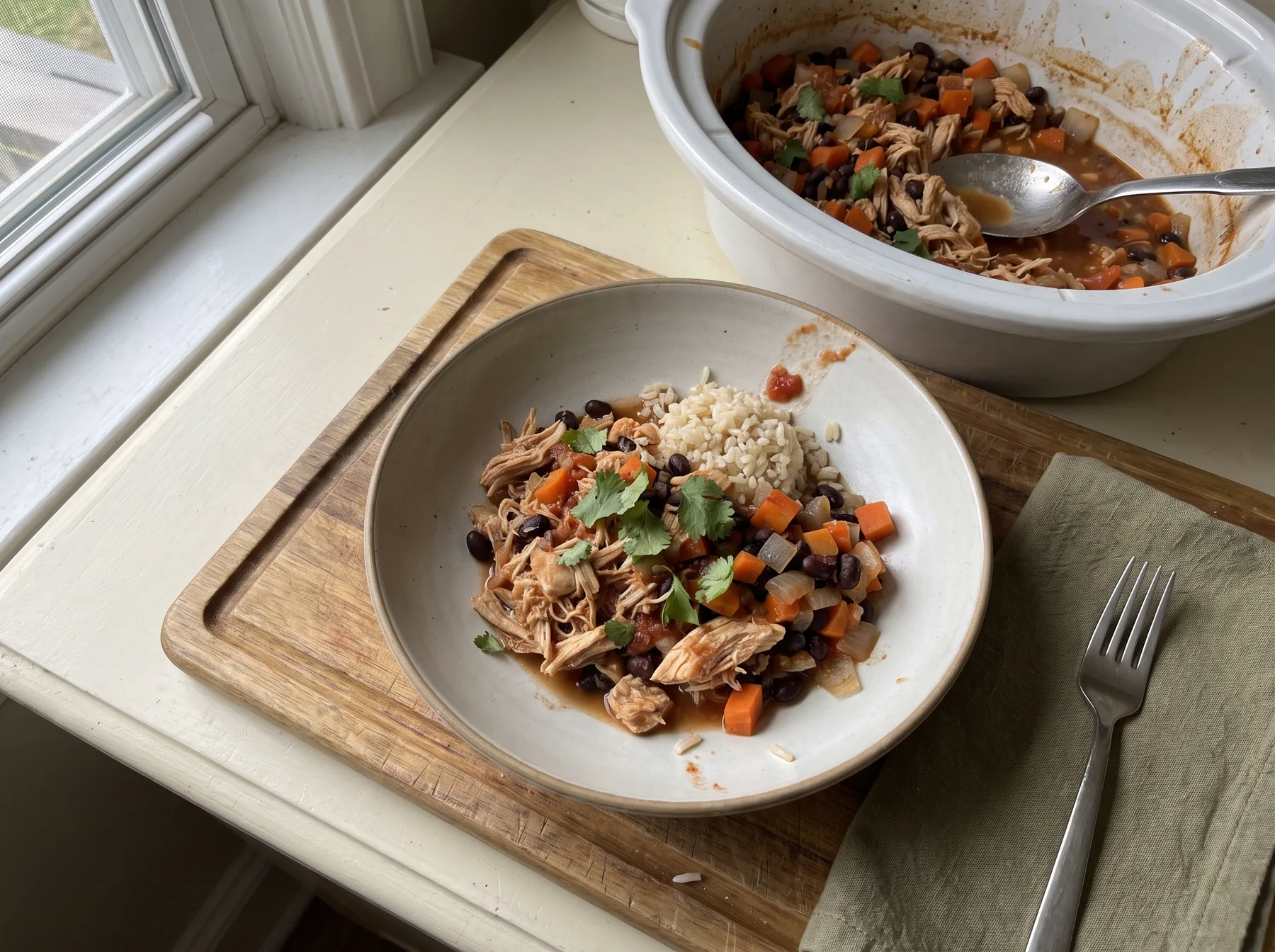 Bowl of shredded chicken, rice, beans, and carrots beside a slow cooker near a window.