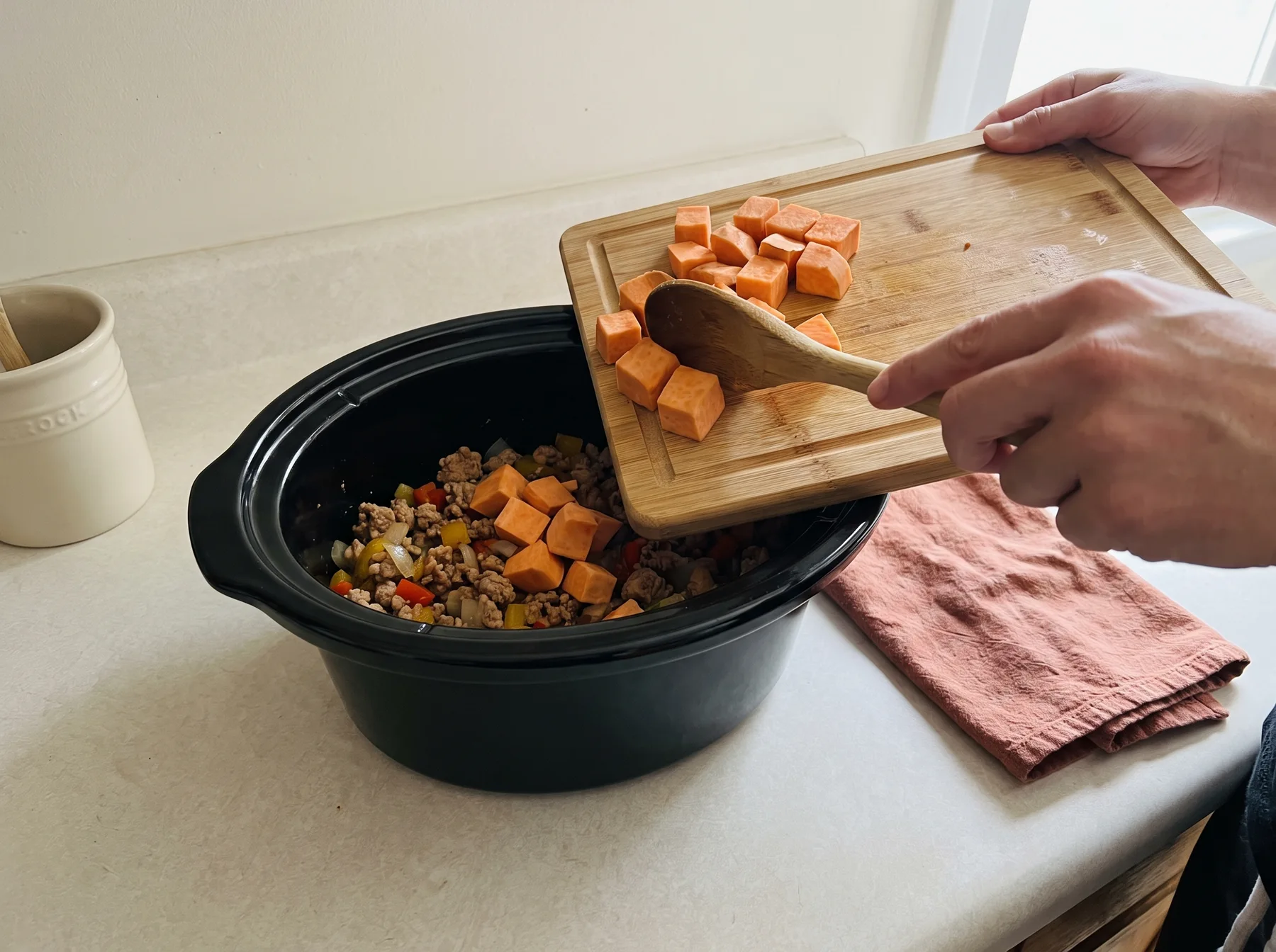 Person adds cubed sweet potatoes to ground turkey and vegetables in a slow cooker.