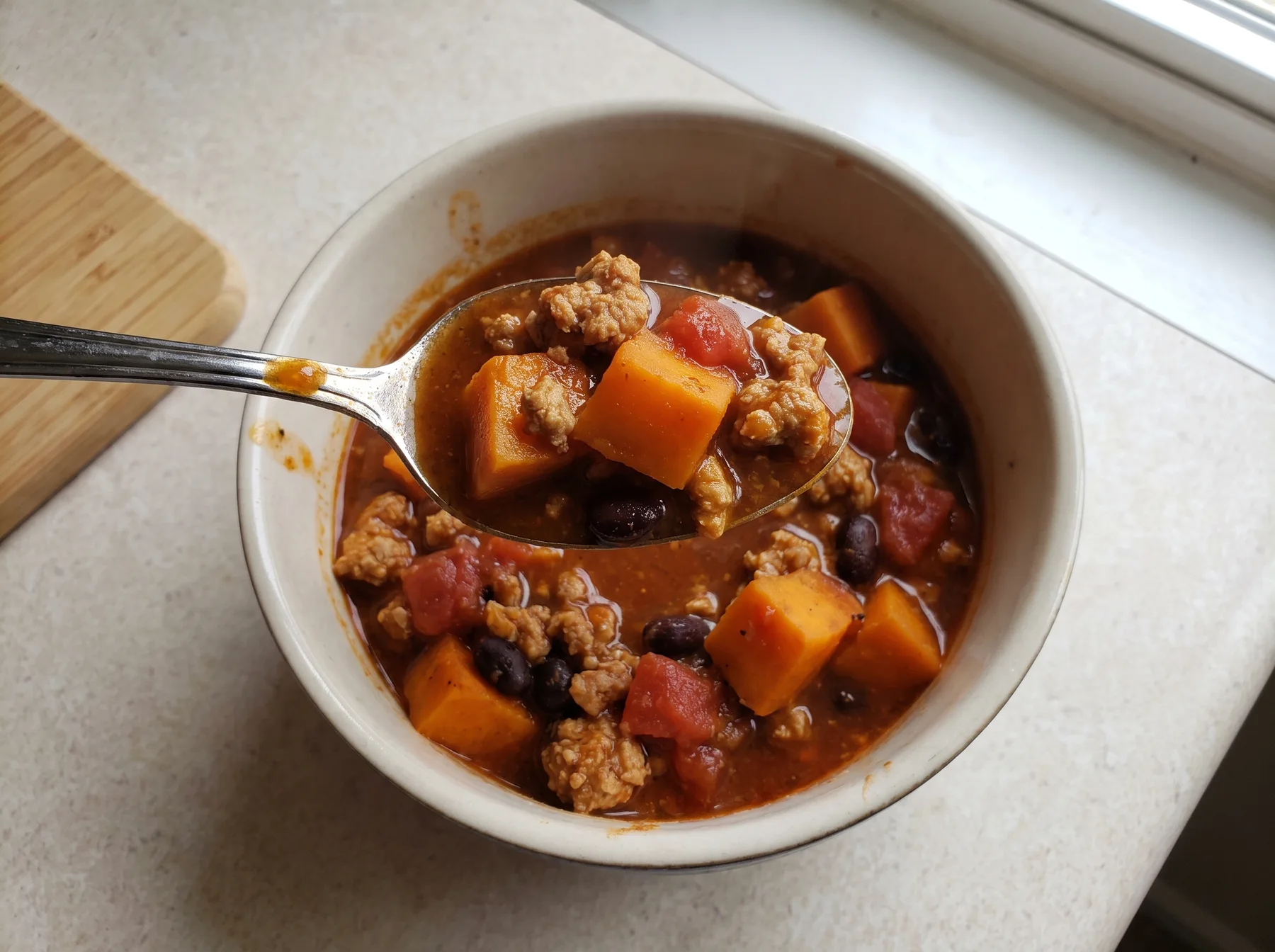 Bowl of turkey sweet potato chili with beans and tomatoes, spoonful lifted beside a cutting board.