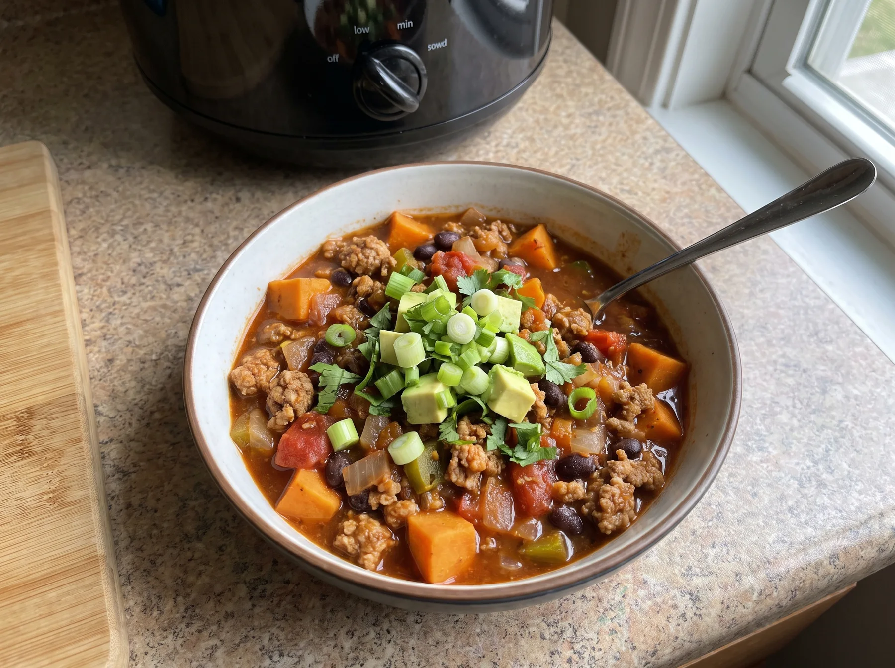Bowl of turkey sweet potato chili topped with avocado and scallions beside a slow cooker.