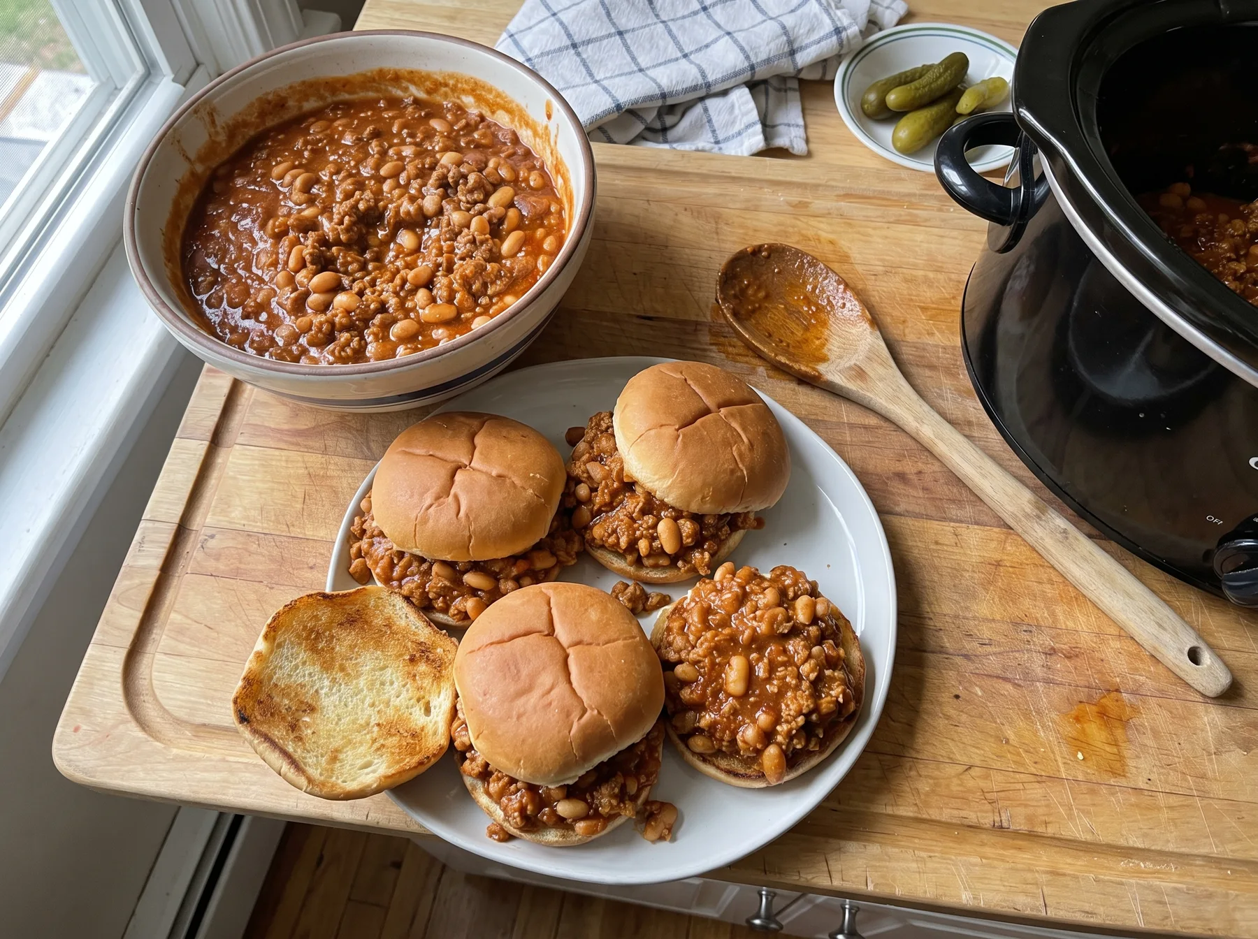 Three turkey and white bean sloppy joes on a plate beside a pot and slow cooker.