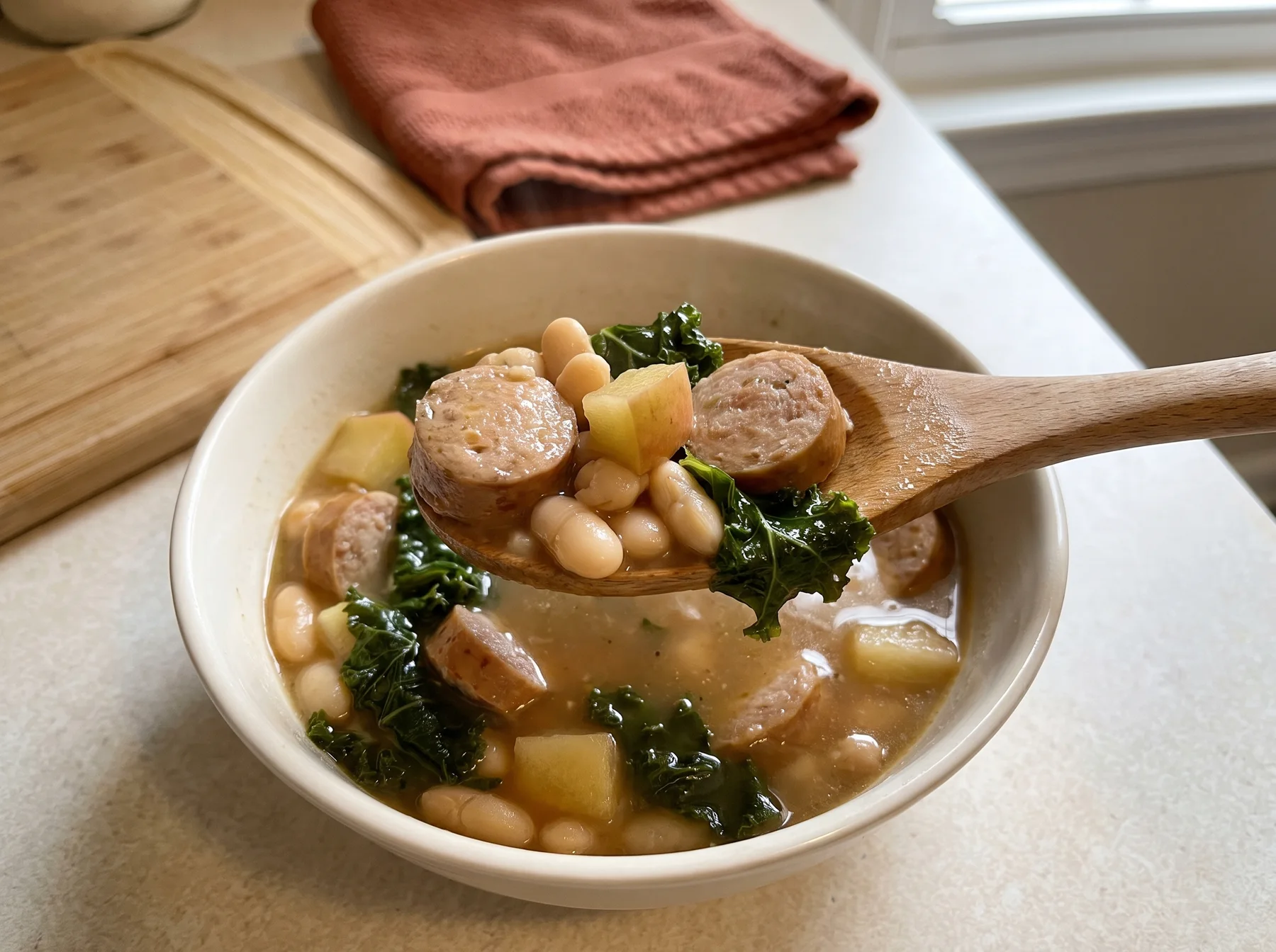 Wooden spoon lifting chicken sausage, white beans, potatoes, and greens from a bowl of stew.