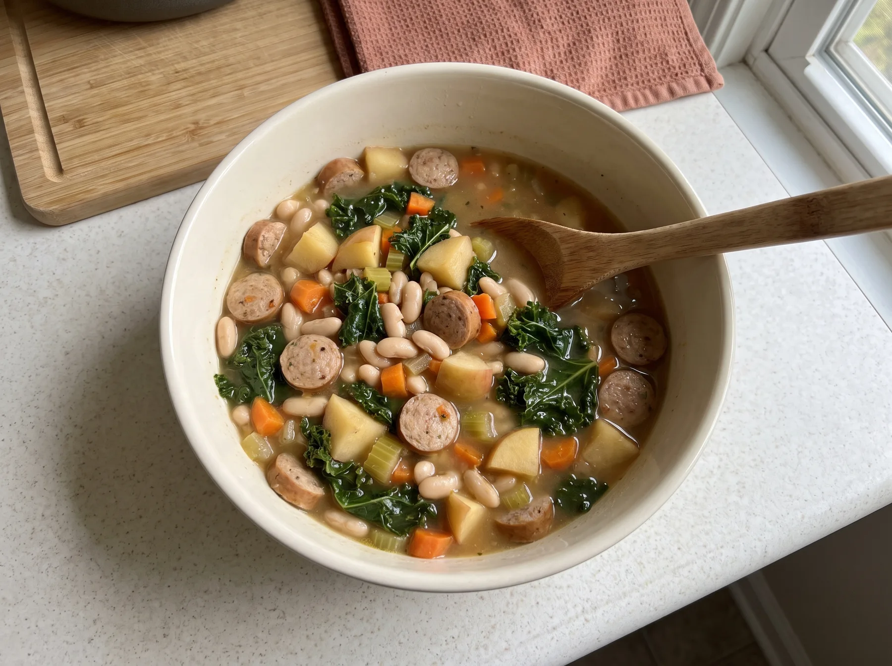 Bowl of chicken sausage and white bean stew with kale, potatoes, and a wooden spoon.