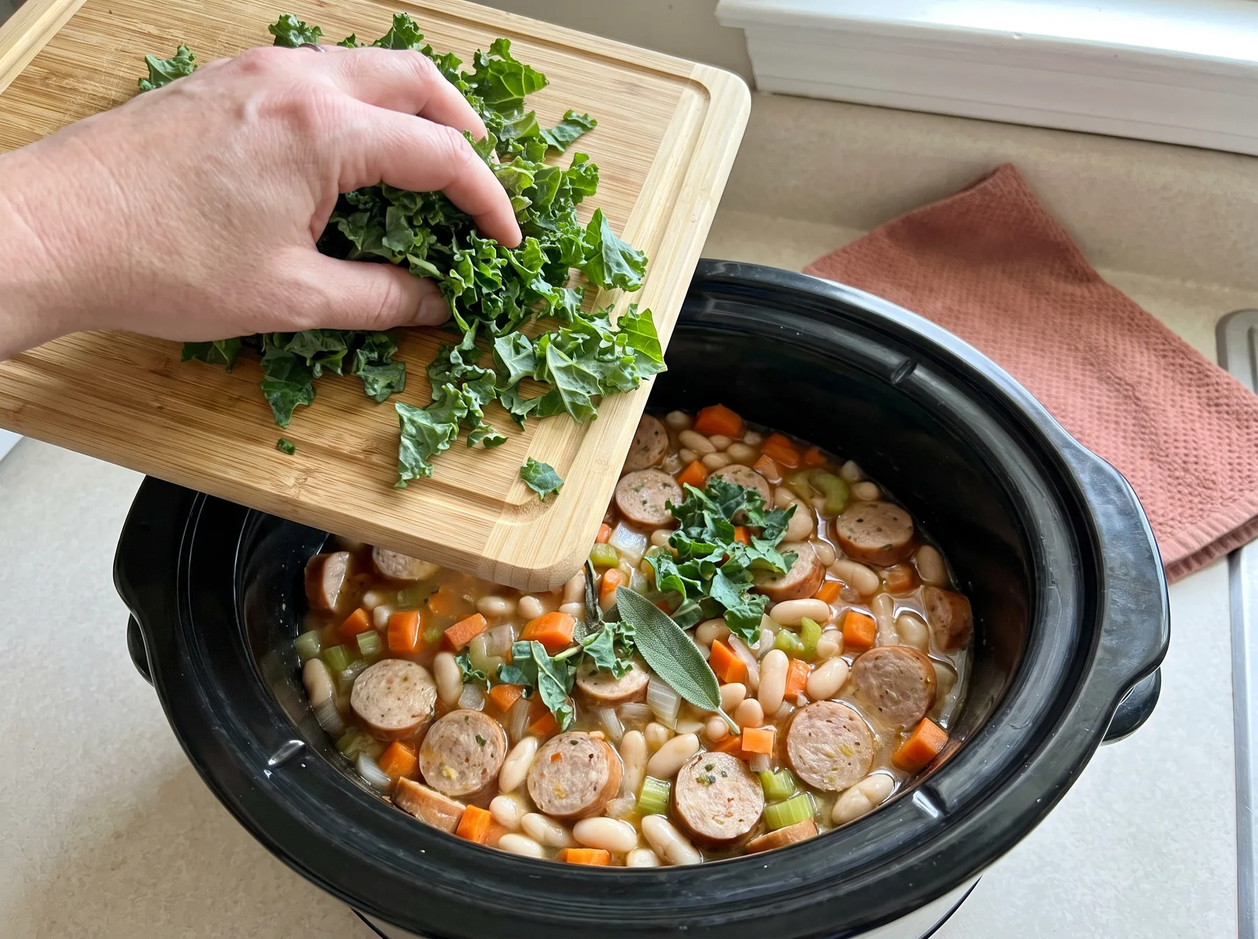 Hand adds chopped kale to slow cooker with sliced sausage, white beans, and vegetables.